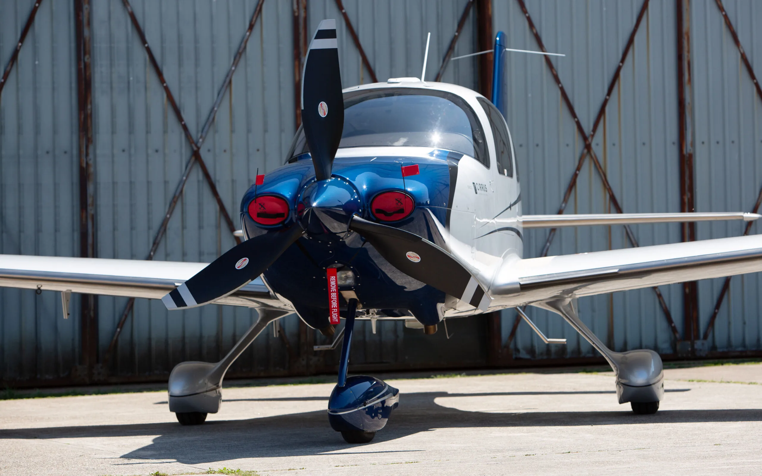 A front view of a blue and white Cirrus SR22 aircraft with a propeller, parked in front of a hangar.