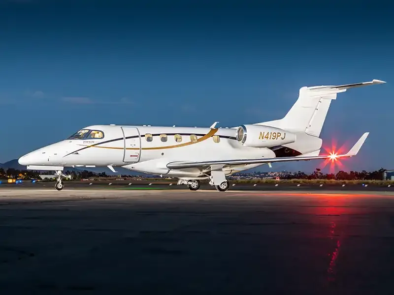 A white Embraer Phenom 300 business jet with gold accents is parked on a tarmac at dusk, with its navigation lights on.