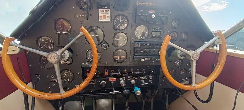The cockpit of a vintage aircraft, showing the instrument panel with various gauges and two wooden control yokes.