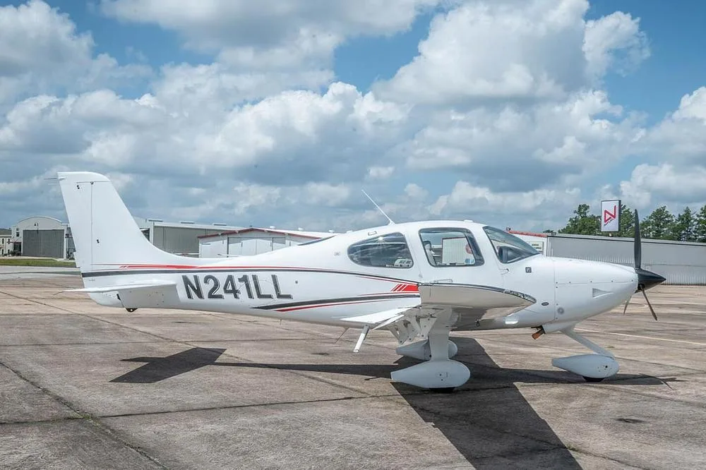 A side view of a white Cirrus SR22 aircraft, registration N241LL, parked on an airfield with hangars in the background.