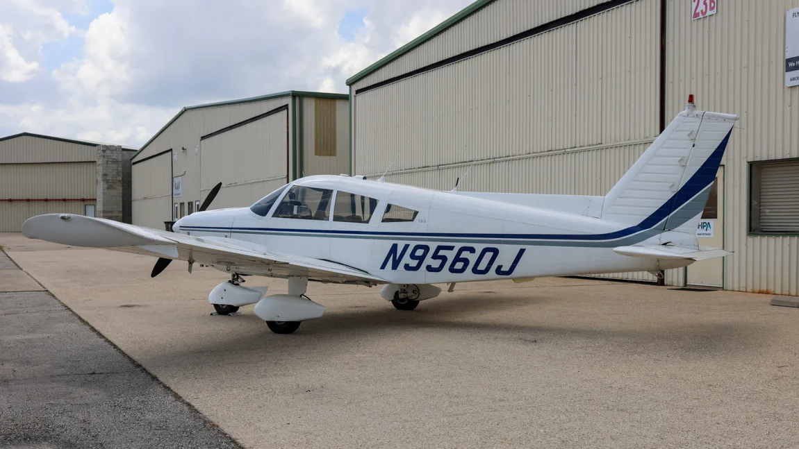 Side view of the white Piper Cherokee 180 (N9560J) parked on an airfield with hangars in the background.