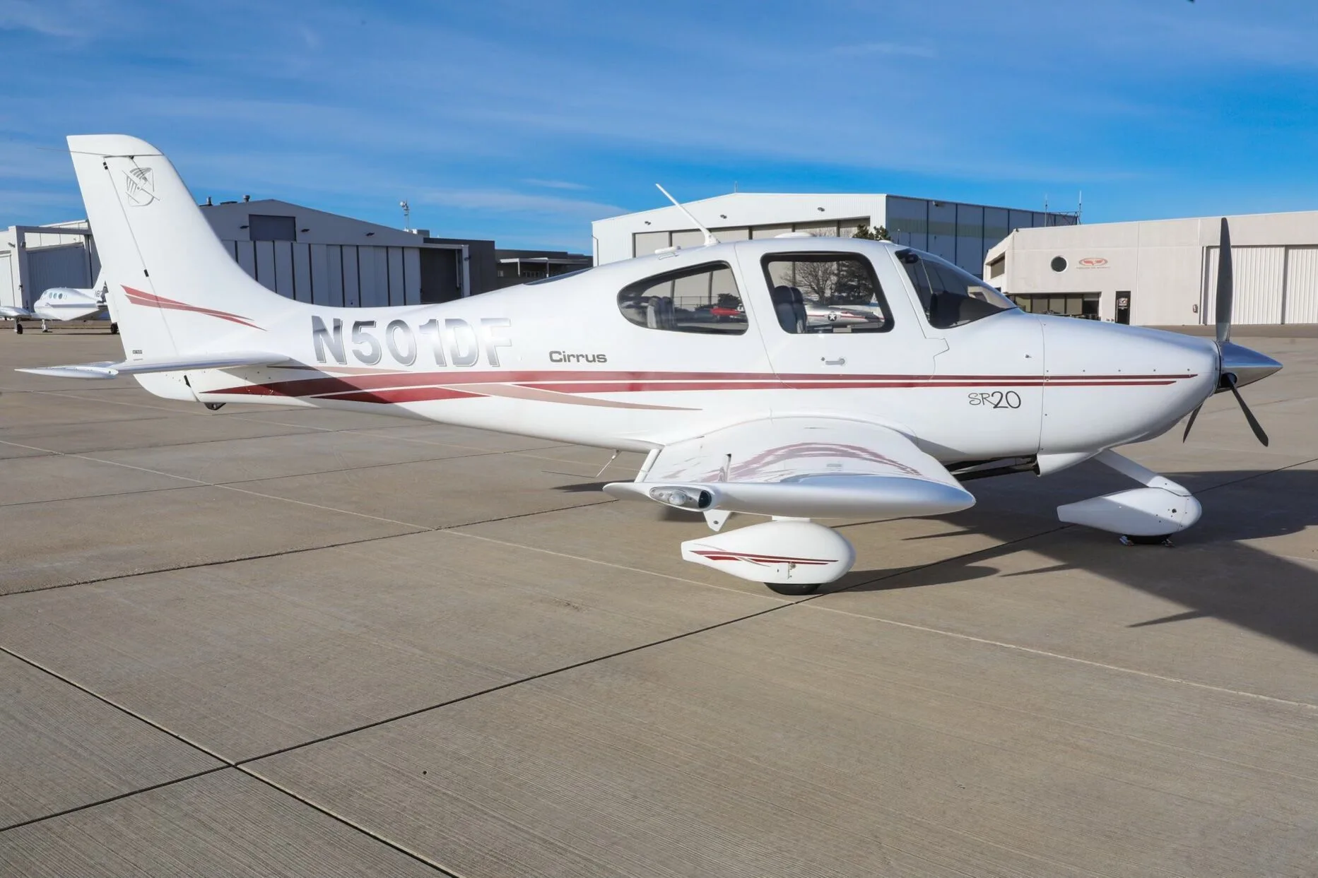 A white Cirrus SR20 aircraft with red stripes, tail number N501DF, parked on an airfield with hangars in the background.