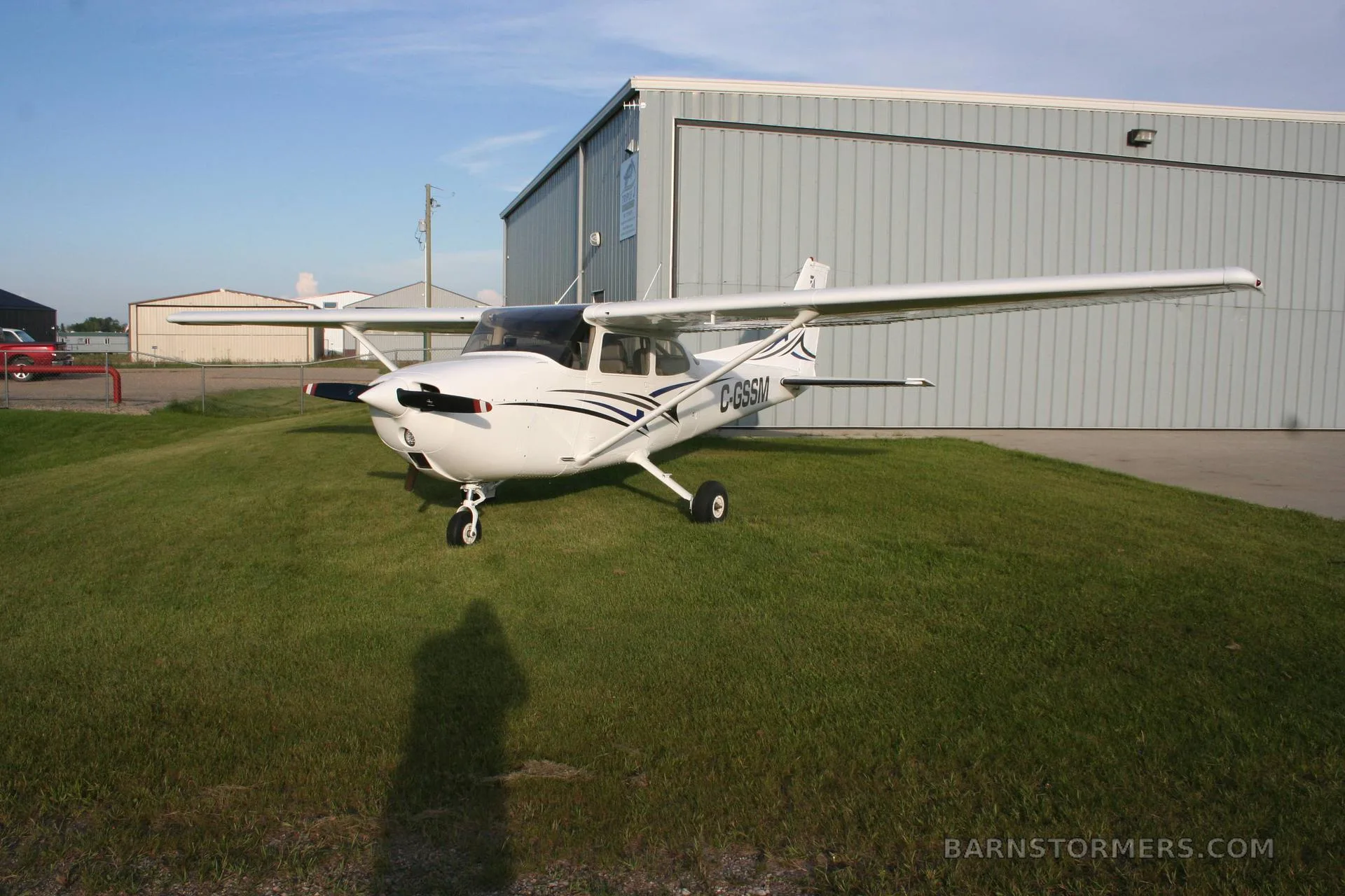 A white Cessna 172 (C-GSSM) with blue and black stripes is parked head-on on green grass in front of a hangar.