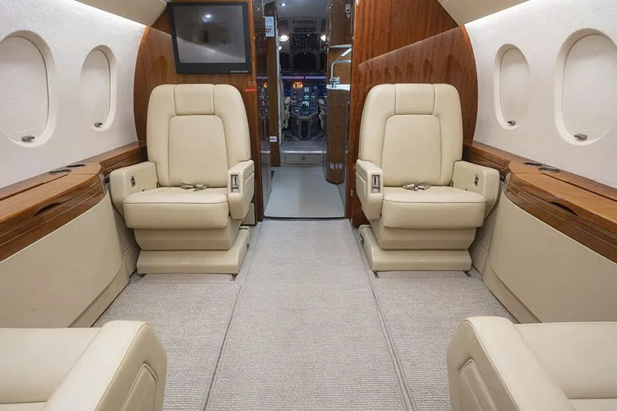 The luxurious interior of a private jet cabin with two cream-colored leather seats facing forward and a view into the cockpit.