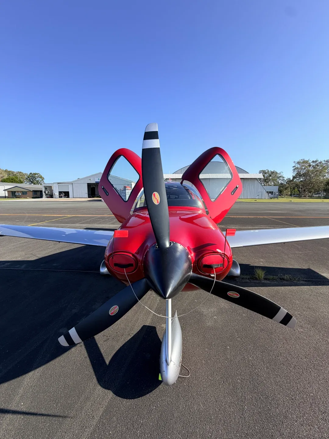 A red single-engine aircraft with open doors is parked on an airfield, viewed from the front.