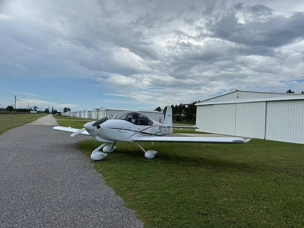 A white Van's RV-10 aircraft with red and black accents, registration N511DL, parked on grass next to a gravel path and hangars.