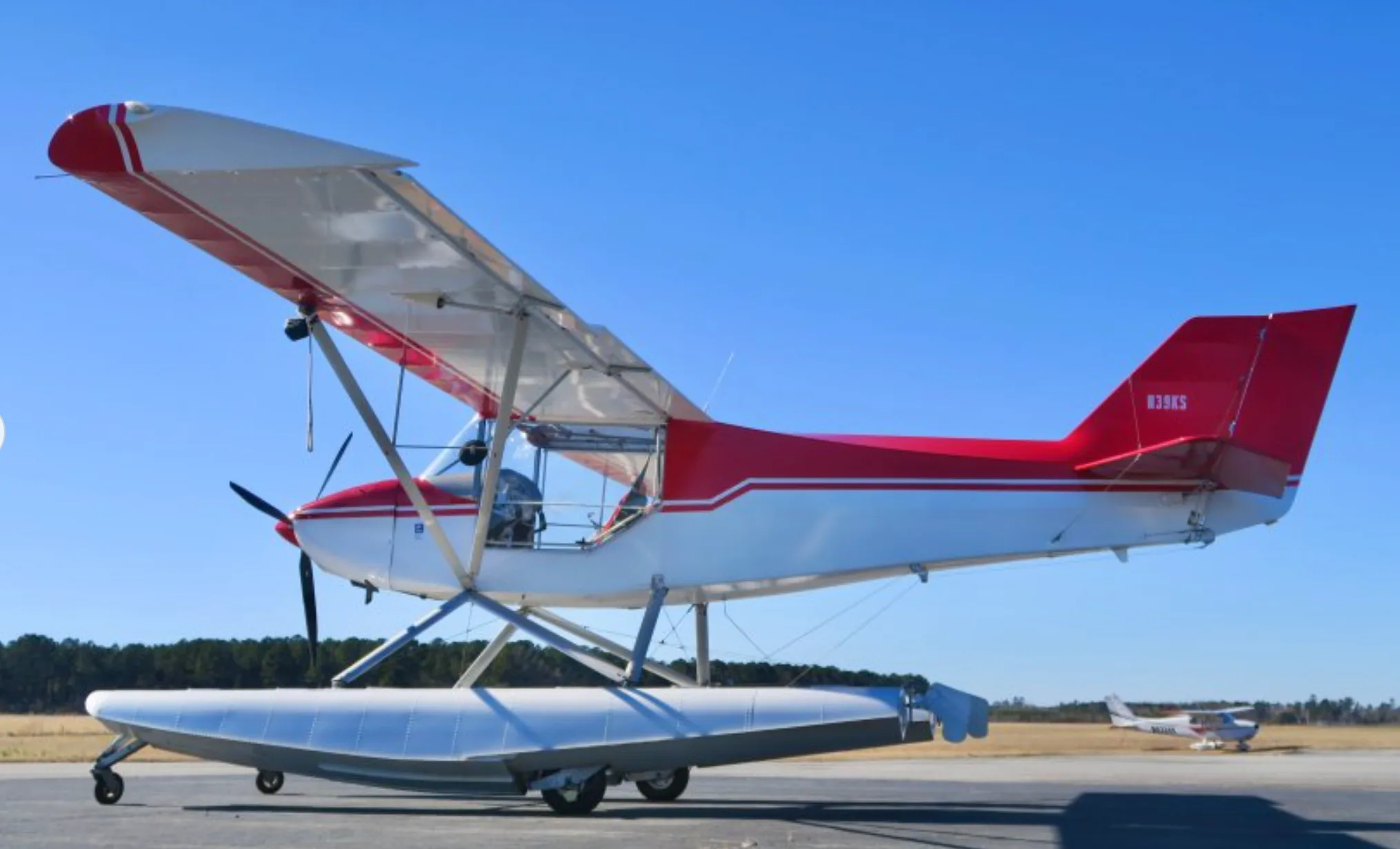 A red and white N39KS floatplane with its wings slightly angled, parked on an airfield with another aircraft in the distance.