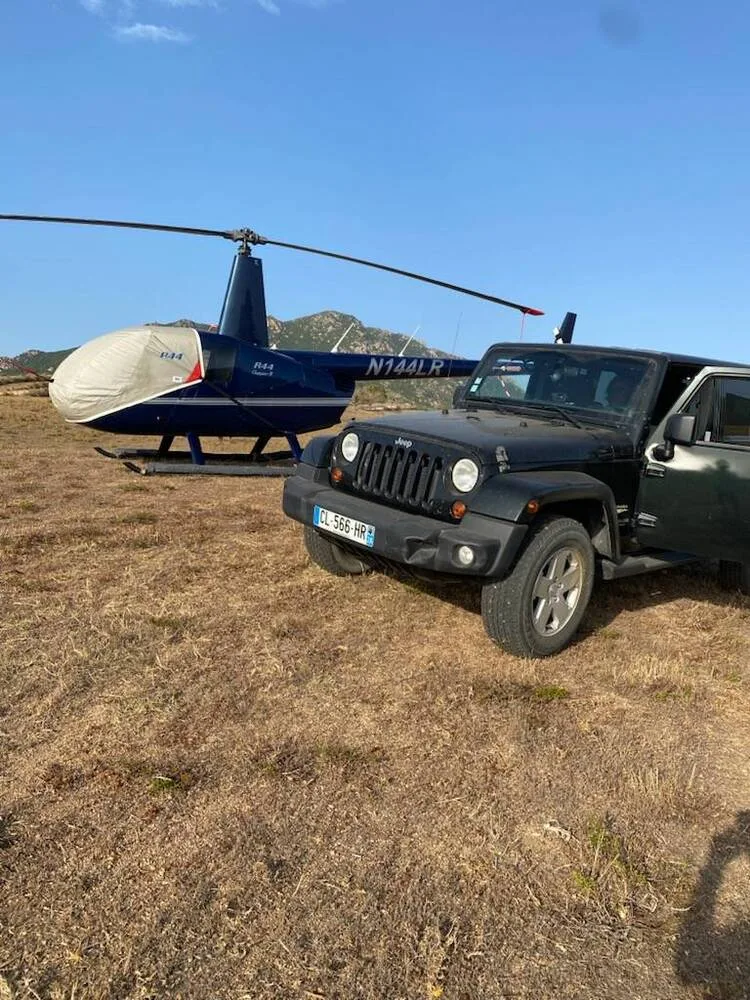 A dark blue Robinson R44 Clipper II helicopter, N144LR, with covered blades, is parked next to a black Jeep Wrangler in a dry field.