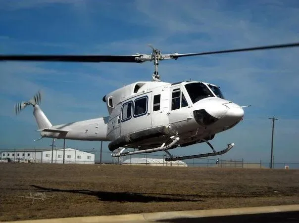 A white Bell 212 helicopter hovers just above the ground in a field under a blue sky.