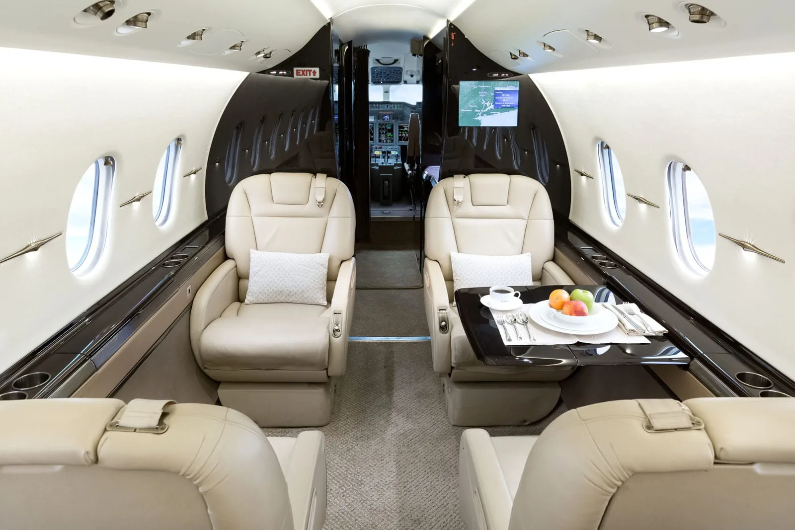 A view down the aisle of a private jet cabin, showing beige leather seats, a table set with fruit and coffee, and a screen displaying a map.