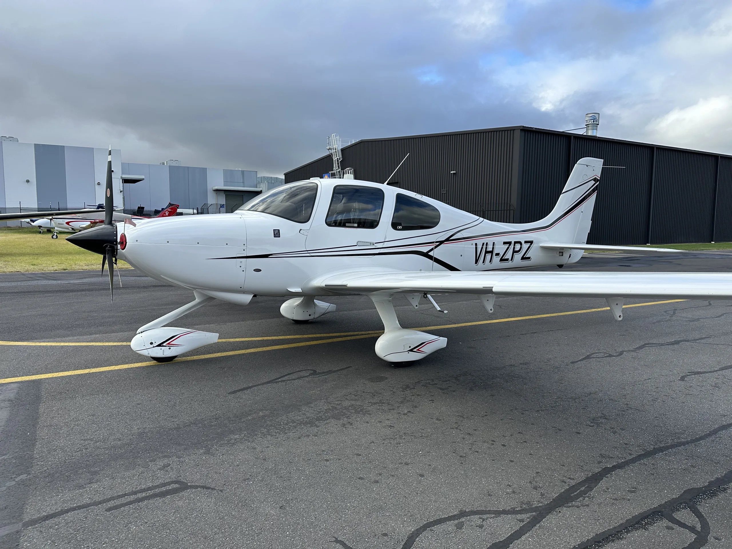 A white single-engine Cirrus SR22 aircraft with black and red stripes parked on an asphalt apron under a cloudy sky.