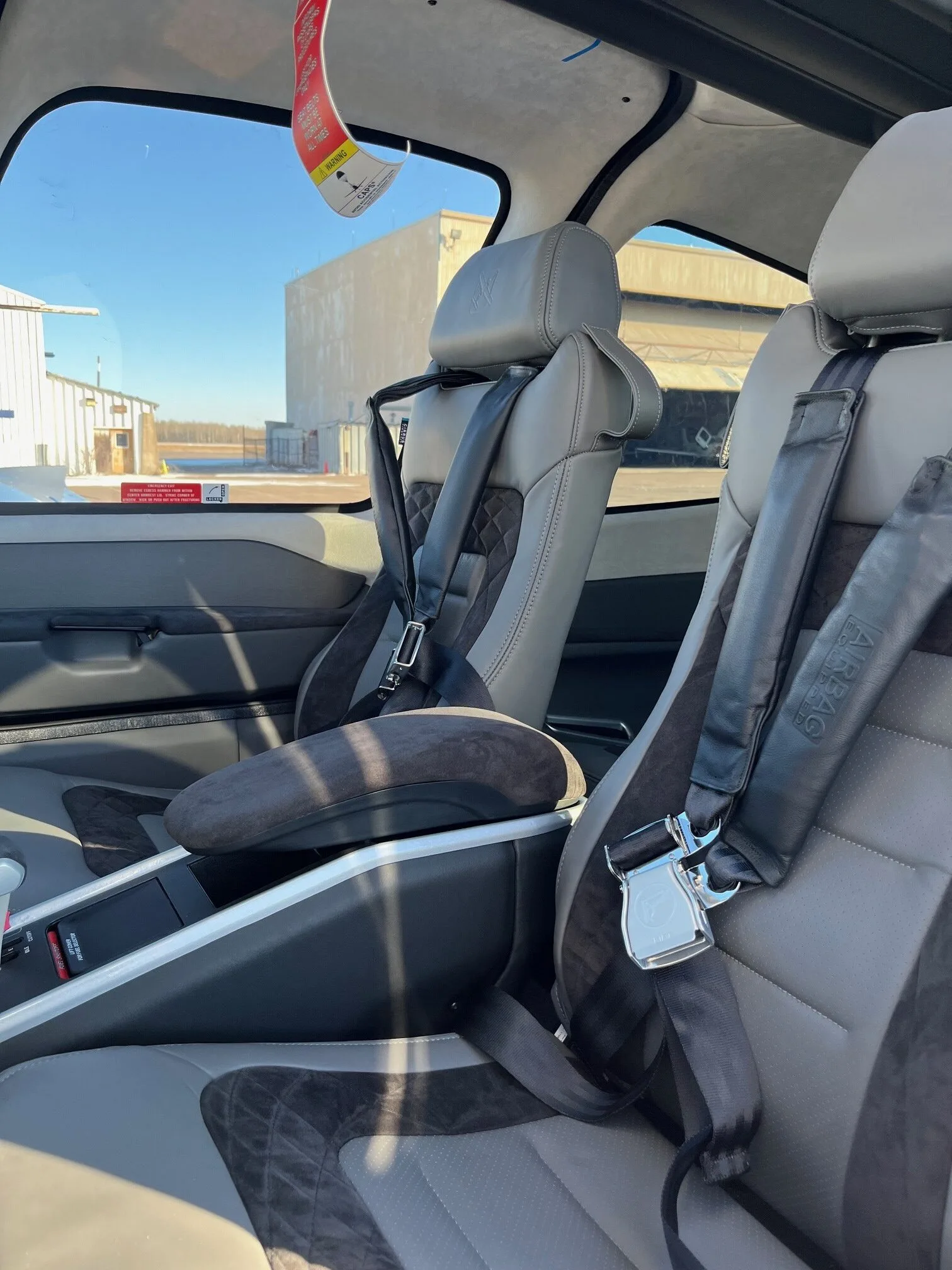 The grey leather interior seats with harnesses inside an aircraft cabin, looking out towards hangars.