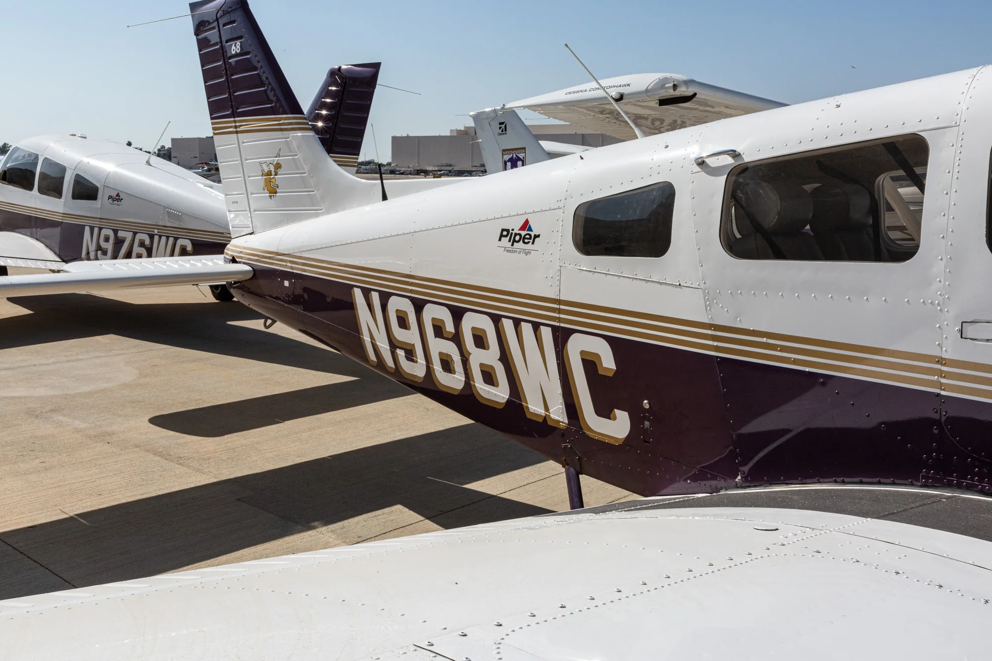 The tail and rear fuselage of a white and purple Piper Archer III aircraft, N968WC, parked on an airfield.