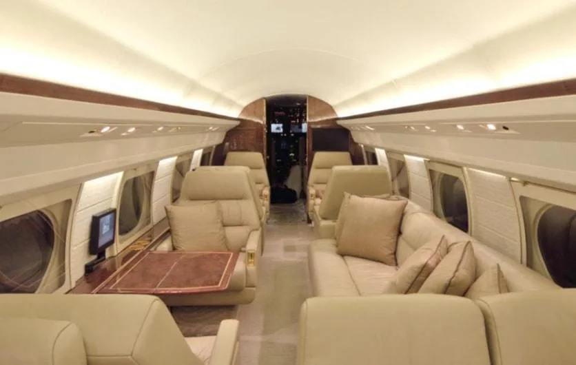 A wide view of a private jet cabin featuring a beige leather sofa on the right and individual seats with a table on the left.