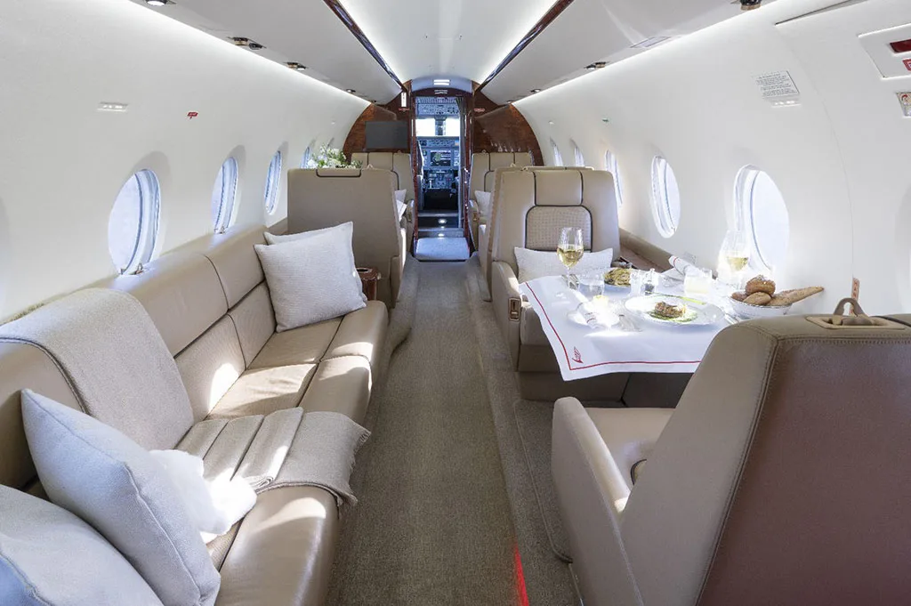 The interior cabin of a private jet with a dining table set for a meal and a long beige leather couch.