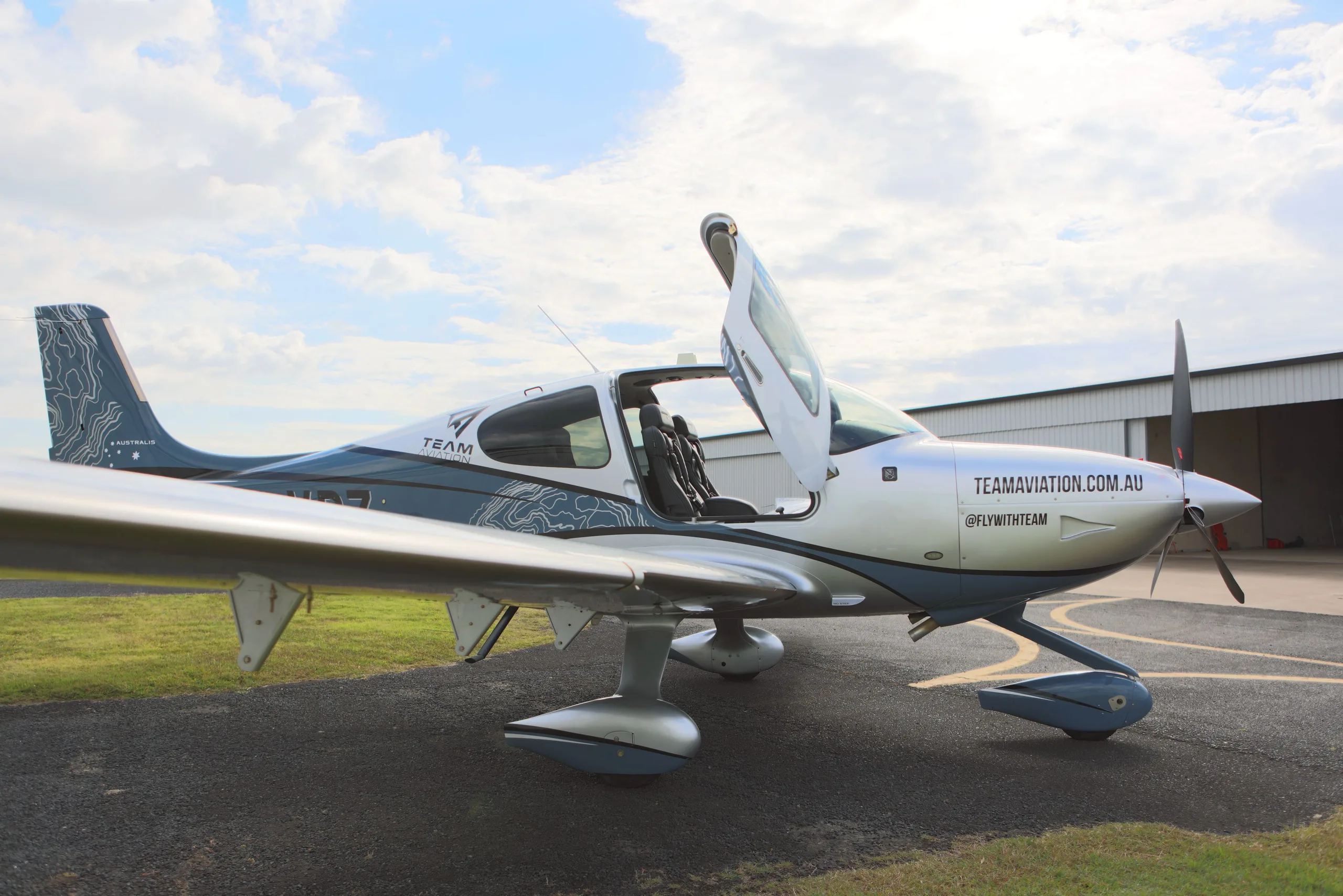 A silver and blue single-engine propeller aircraft with open doors and a topographic map paint scheme is parked on an airfield with a hangar in the background.
