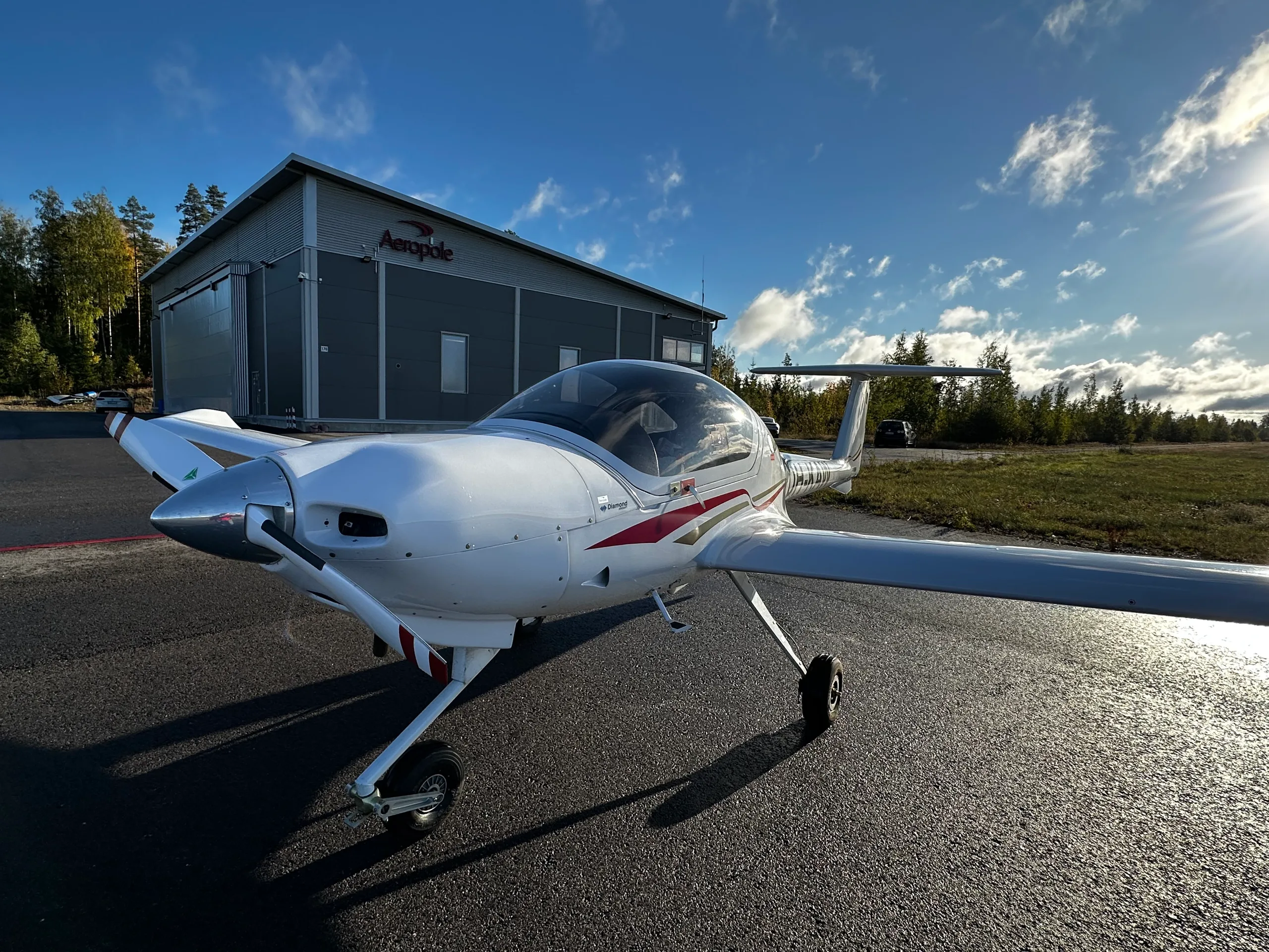 A white Diamond DA20 aircraft with red and black stripes parked on an asphalt apron in front of a hangar under a sunny sky.