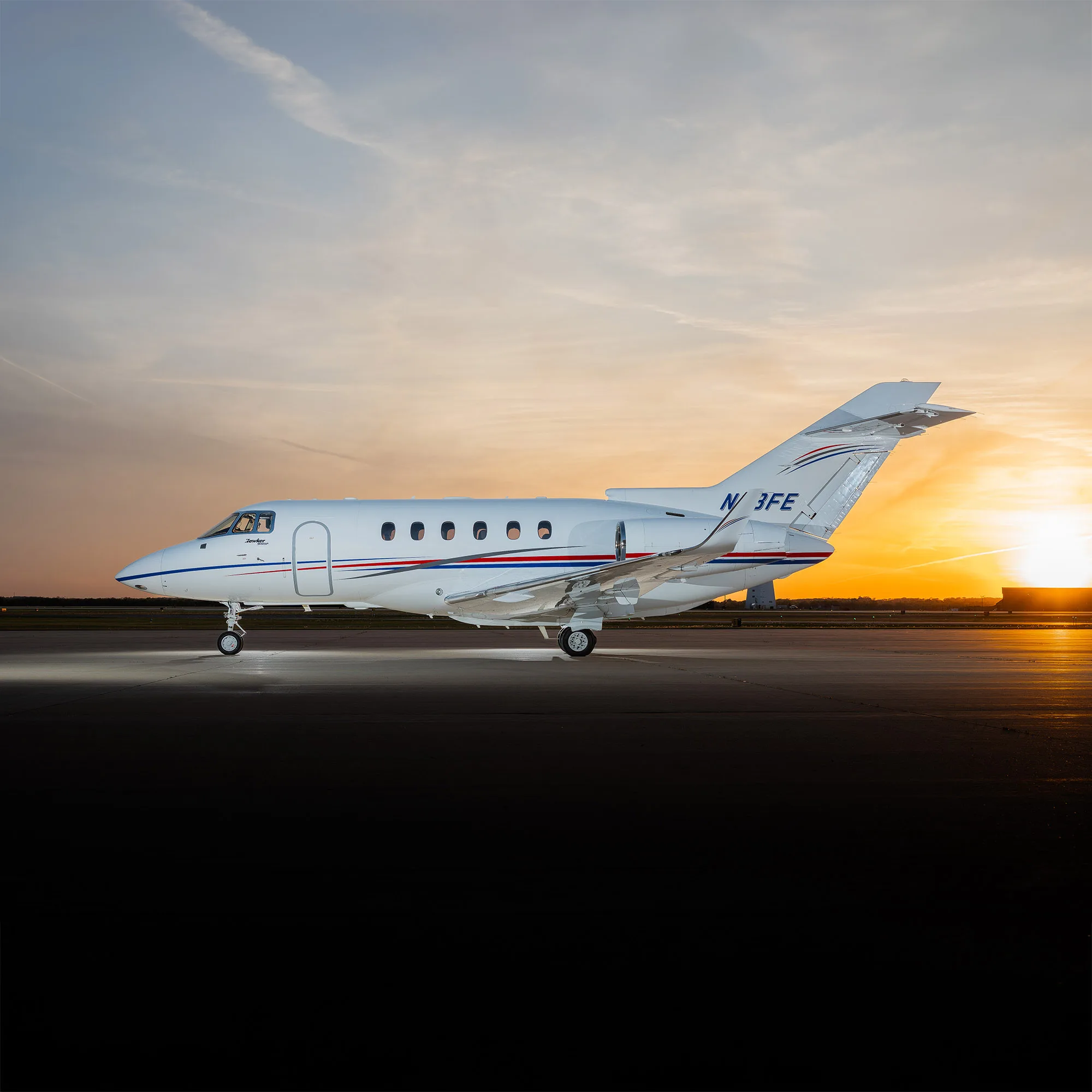 A white business jet with red and blue stripes on the fuselage, registration N13FE, parked on an airfield at sunset.