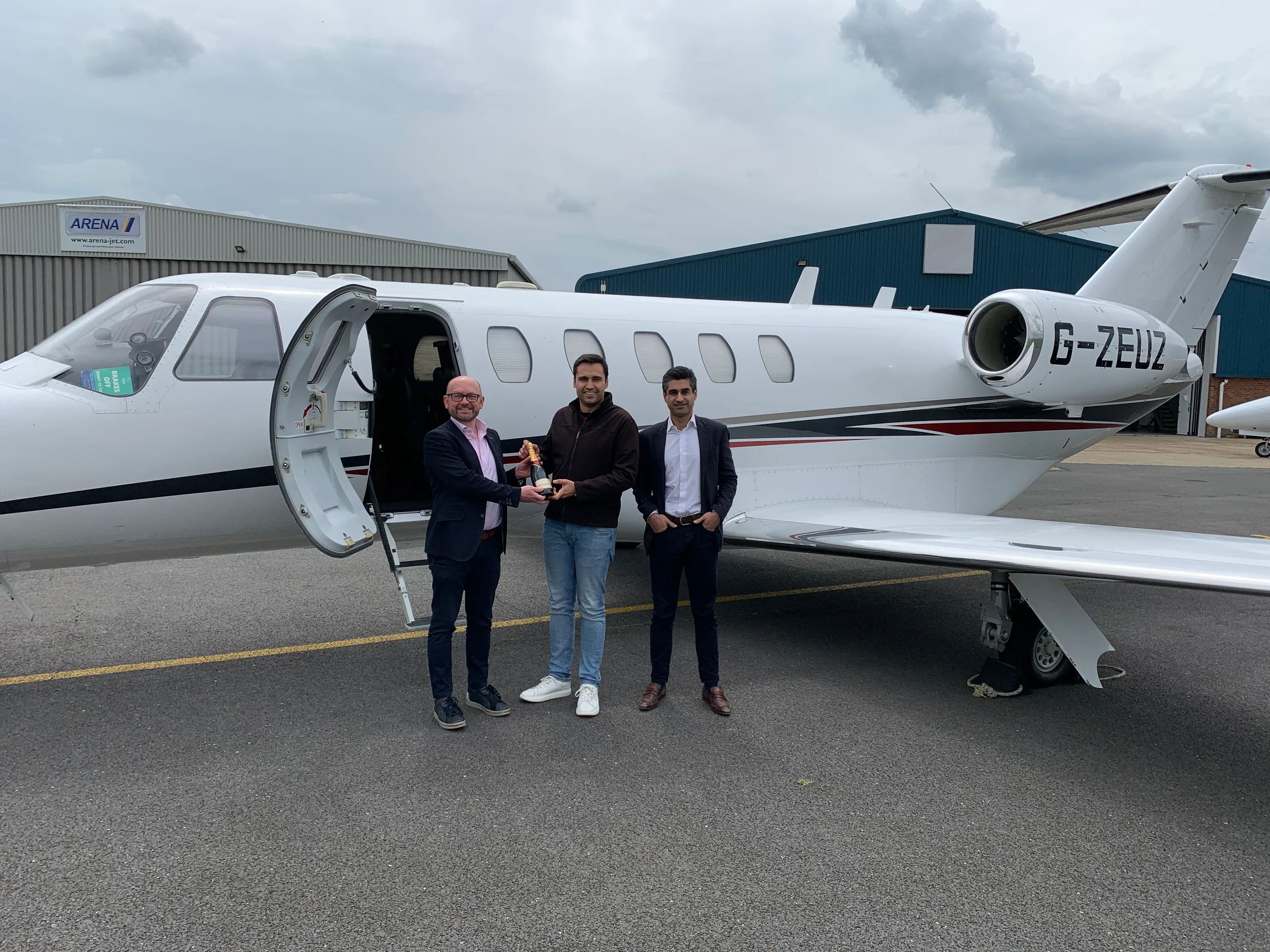 Three men stand next to a white business jet with black, red, and grey stripes, registration G-ZEUZ, on an airport tarmac with a hangar in the background.
