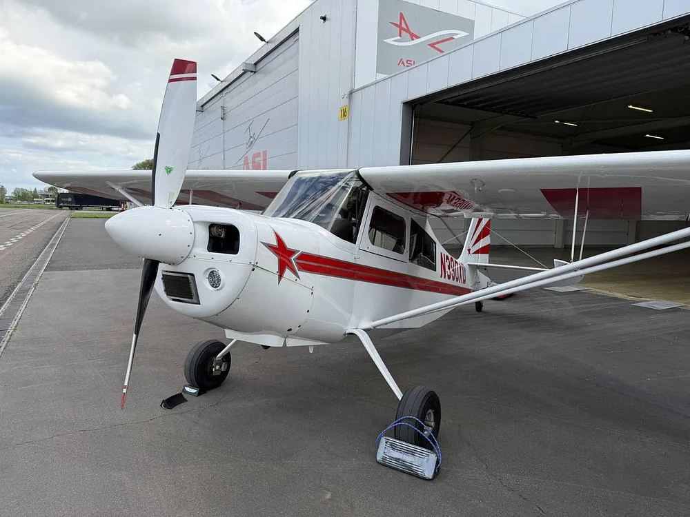 A white single-engine aircraft with red stripes and a red star on the nose, parked on an asphalt apron outside a hangar.