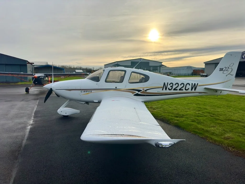 A side view of a white Cirrus SR22 aircraft with N322CW registration, parked on a wet tarmac with the sun low in the sky.