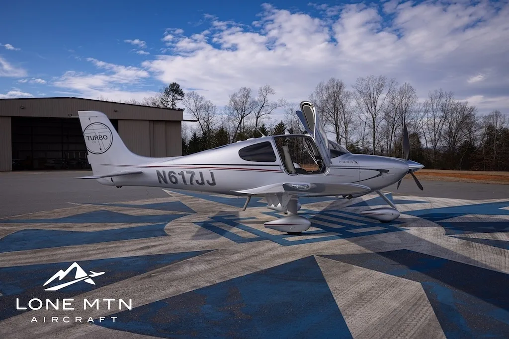 A silver Cirrus SR22T aircraft, N617JJ, with its doors open, parked on an airfield next to a hangar.