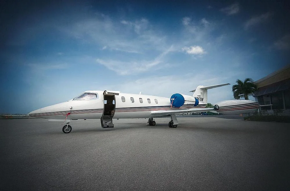 A white Learjet with red and blue stripes, registration N747BW, parked on an airfield under a partly cloudy sky.