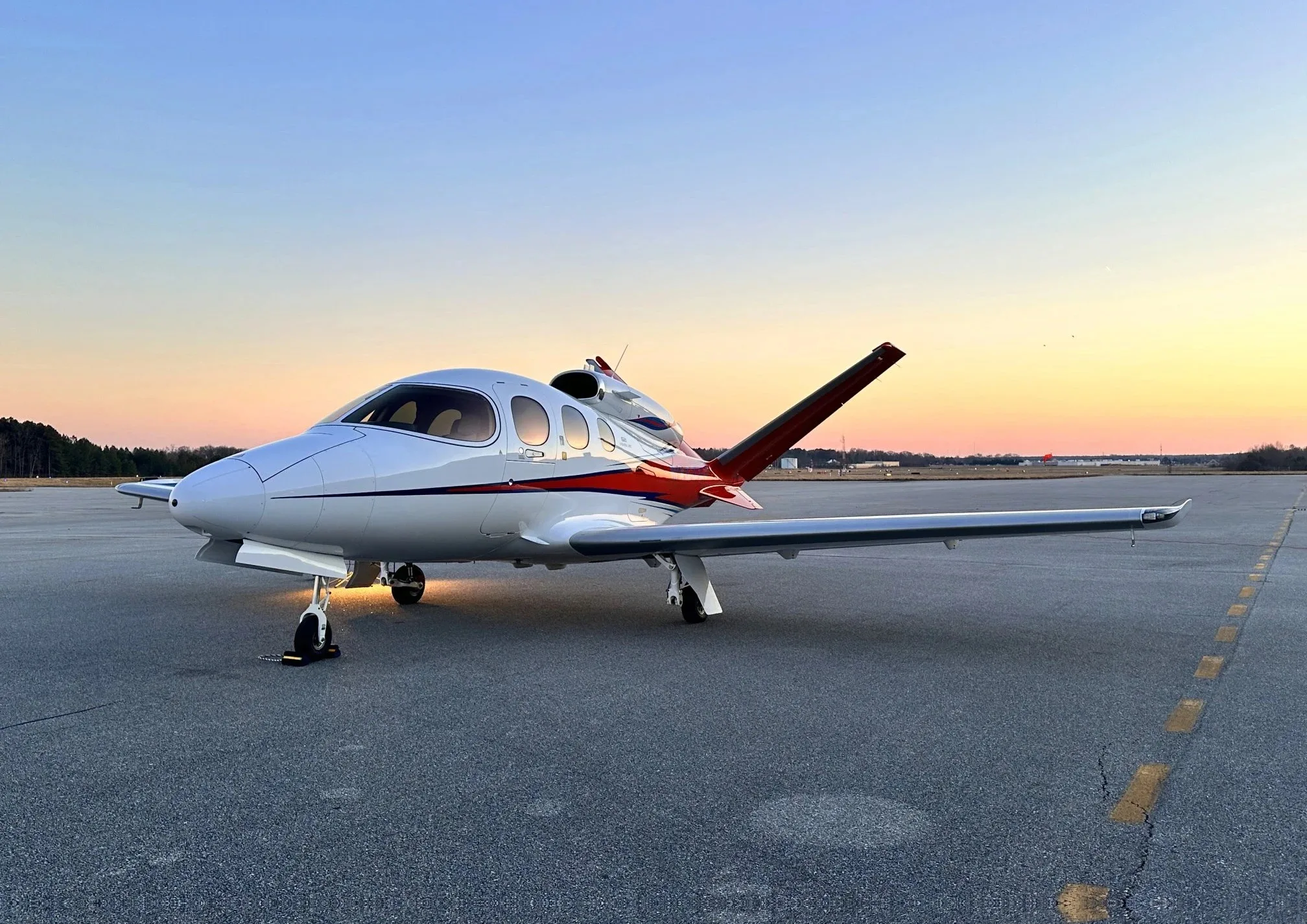 A white Cirrus Vision Jet with red and black accents parked on an airfield at sunset.