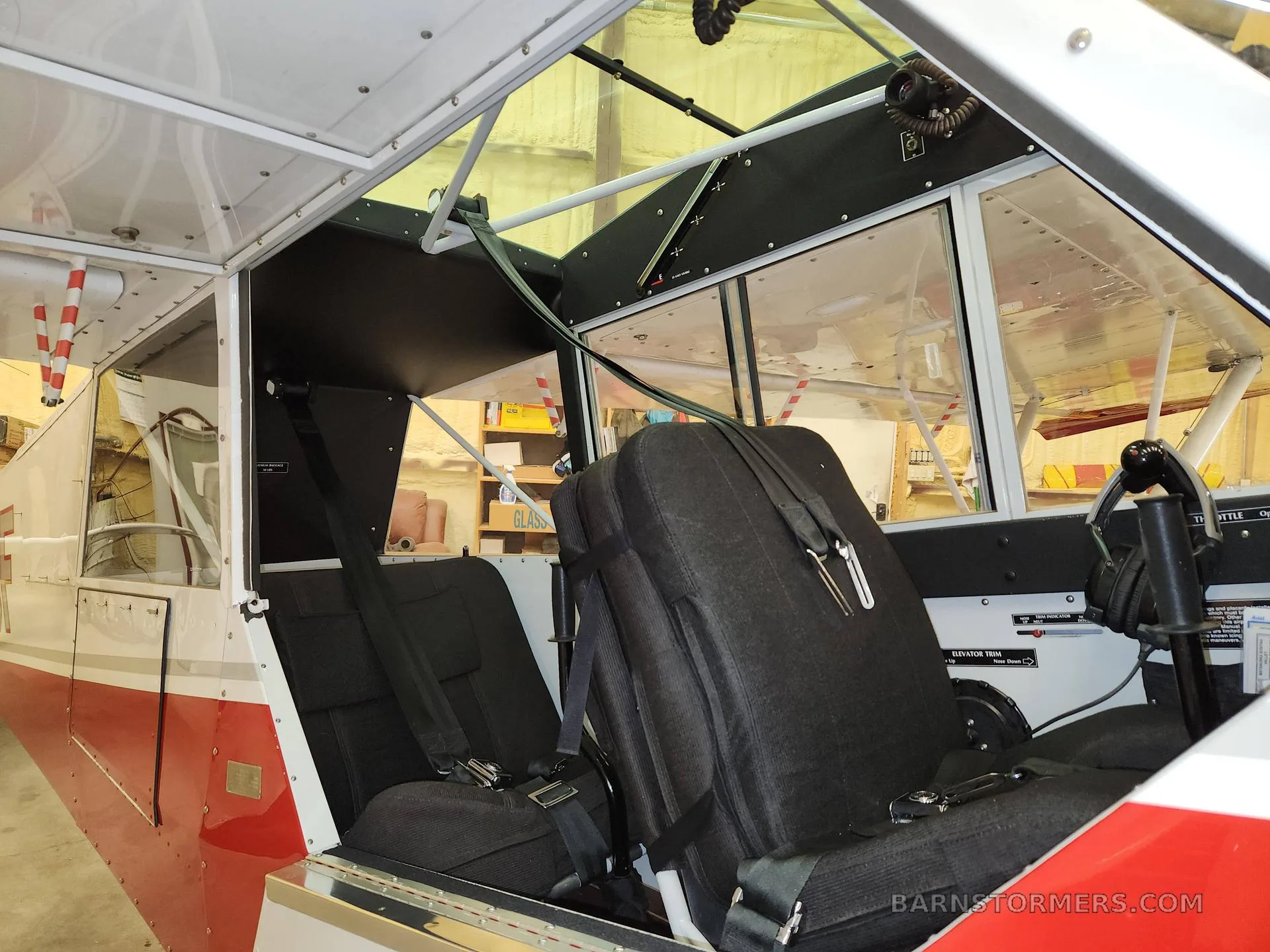 The interior cabin of a red and white aircraft, showing two black seats with harnesses and a view through the windows.
