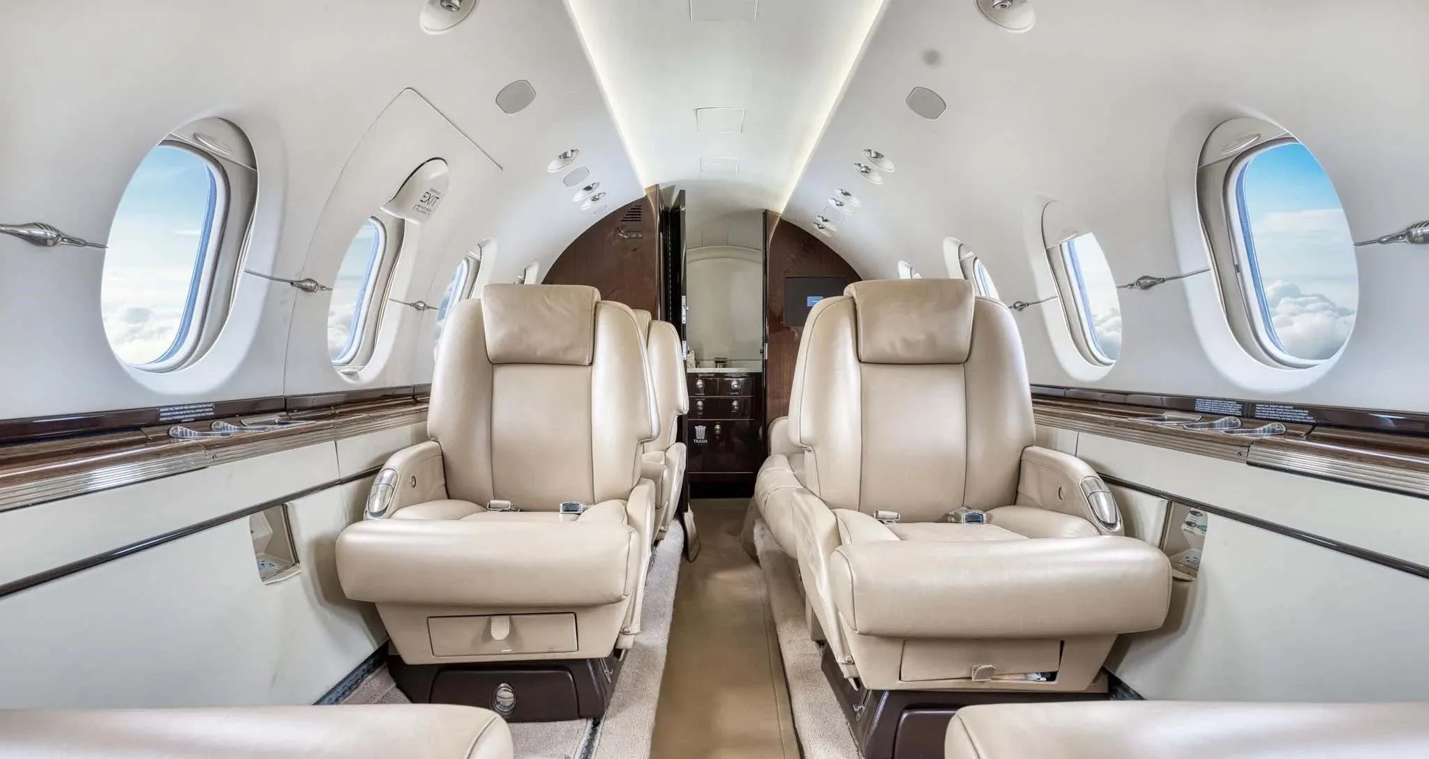 The luxurious interior of a private jet cabin featuring light beige leather seats and dark wood paneling.