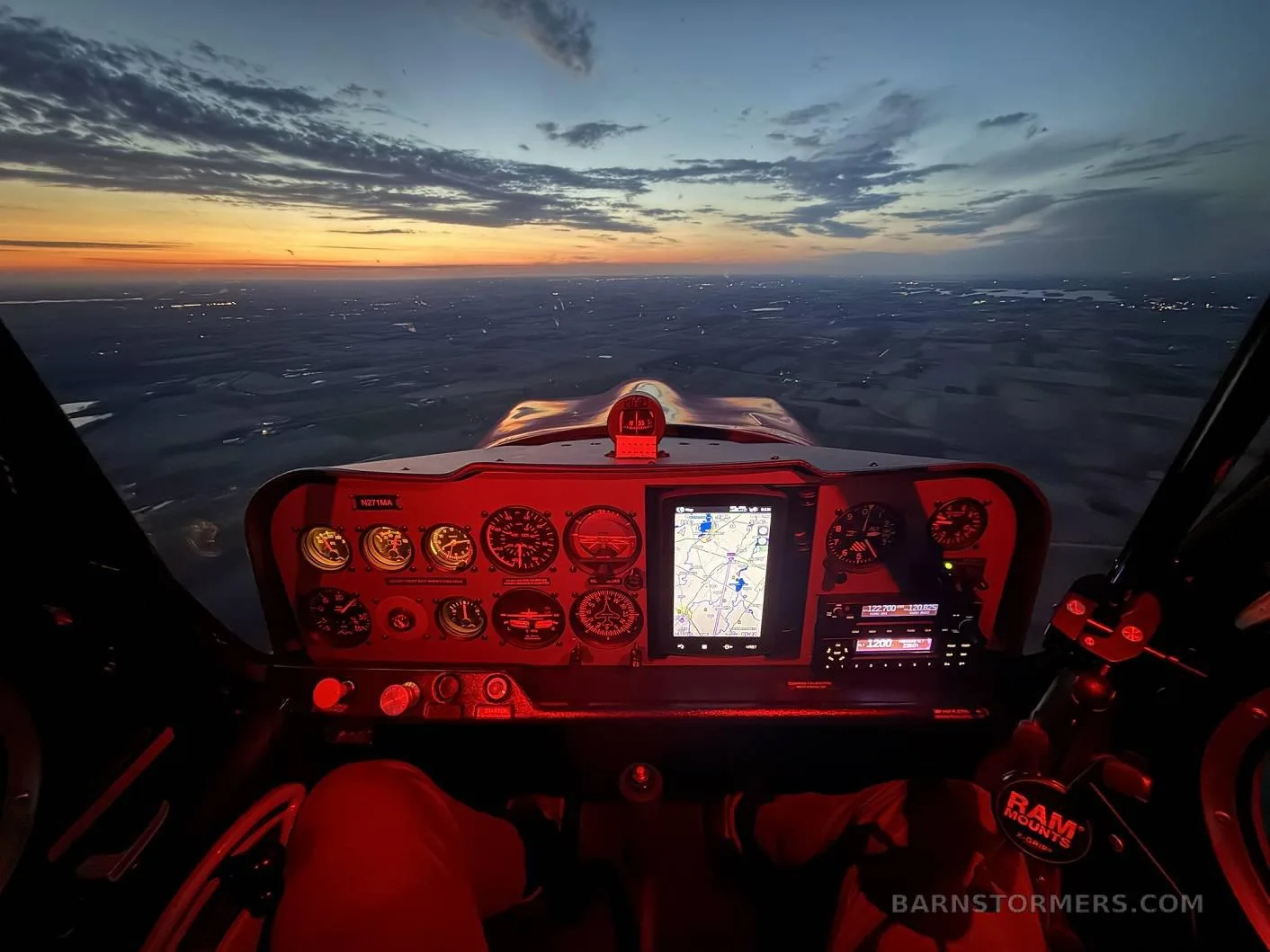 A pilot's view from the cockpit at dusk, with the instrument panel illuminated in red and city lights visible below.