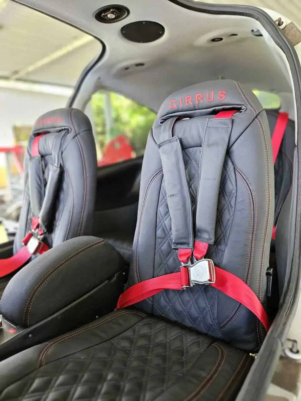The front black leather seats with red stitching and "CIRRUS" embroidery, featuring red seatbelts, inside an aircraft cabin.