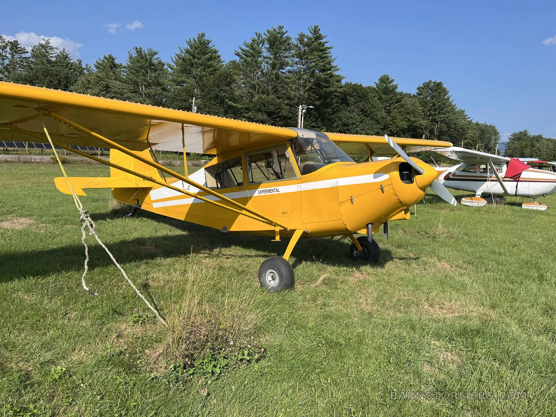 A yellow high-wing experimental aircraft with white stripes parked on a grassy field under a blue sky.