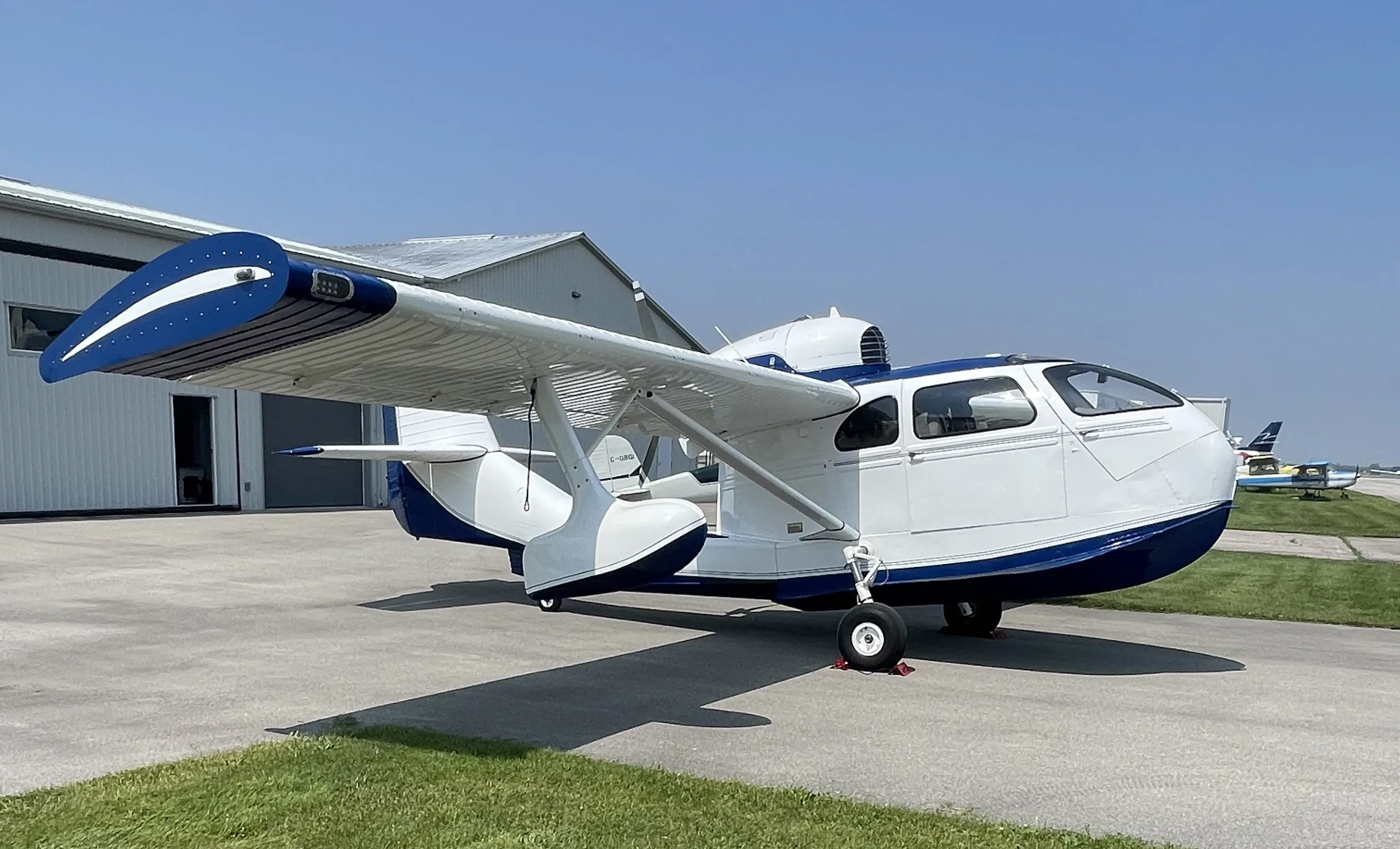 A white and blue amphibious aircraft is parked on a tarmac with hangars and other planes in the background under a clear sky.