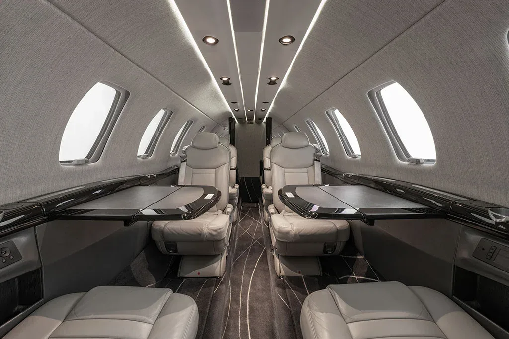 The interior cabin of a private jet shows light gray leather seats with black fold-out tables extended.