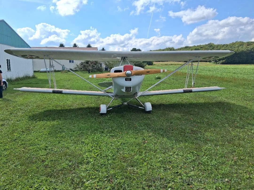 A white biplane with a red nose cone and wooden propeller, viewed from the front, parked on green grass under a blue sky.