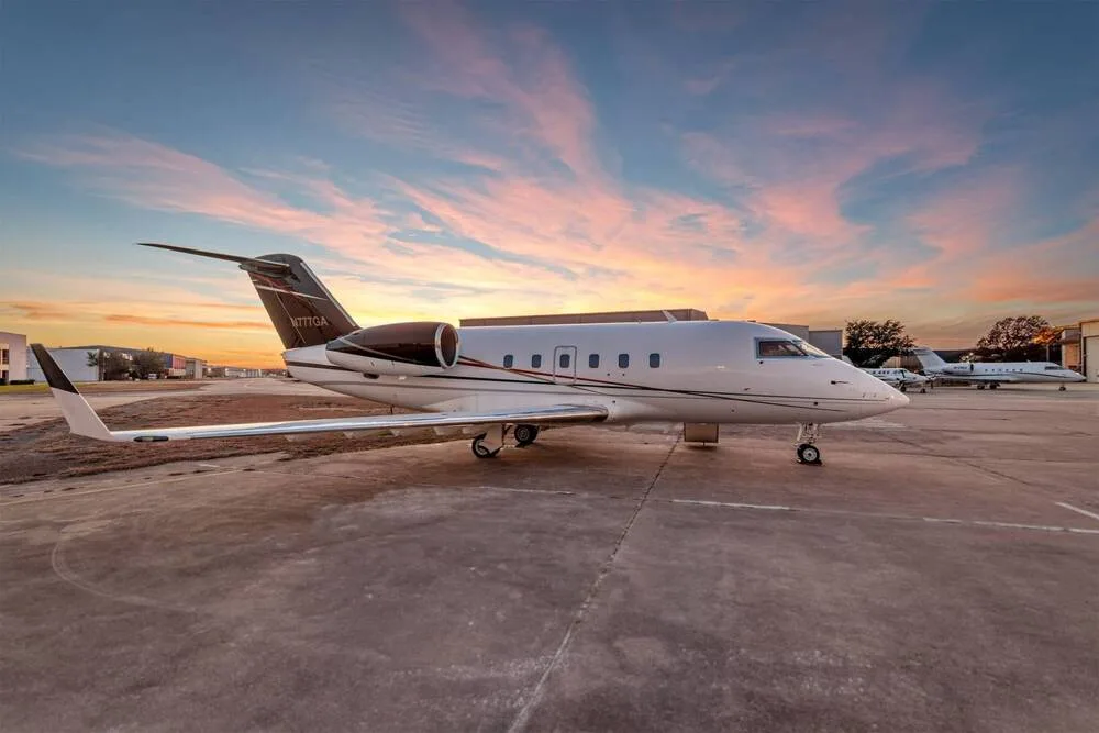 A white business jet with a dark brown and red tail, registration N777GA, parked on an airfield at sunset.
