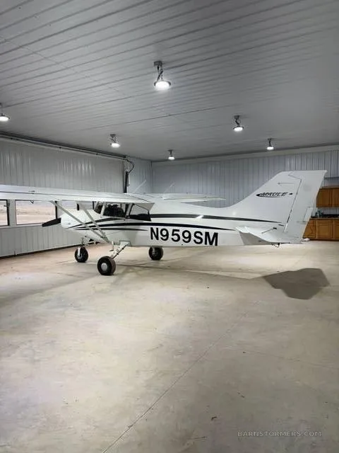 A white Maule M-7-235B aircraft with black stripes and registration N959SM is parked inside a hangar, viewed from the rear-left.