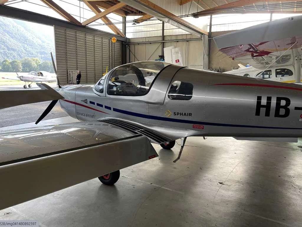 A silver Sonaca aircraft with registration HB-KGF parked in a hangar, viewed from the front-left side.