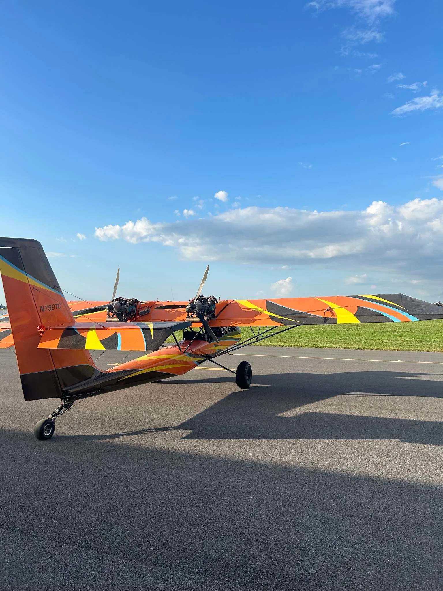 A rear-side view of an orange and black ultralight aircraft with twin engines and a colorful paint scheme, parked on an airfield.
