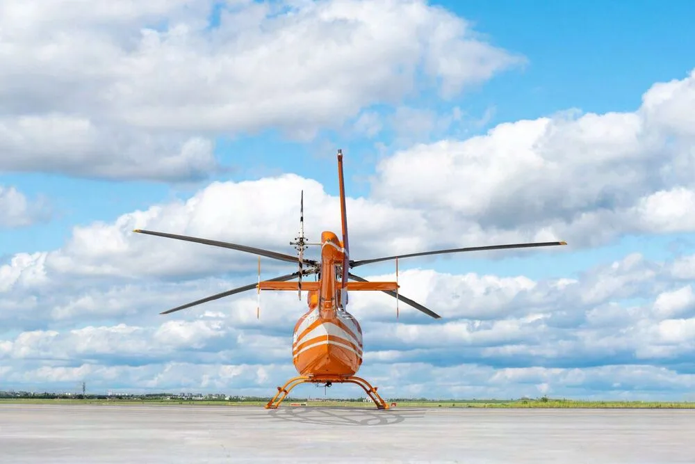 The rear view of an orange Bell 407EXP helicopter with registration B-704L parked on an airfield under a cloudy sky.