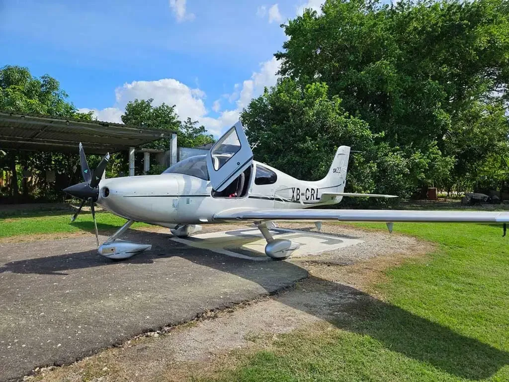 A silver Cirrus SR22 aircraft with its gull-wing door open, parked on a tarmac next to grass and trees under a blue sky.