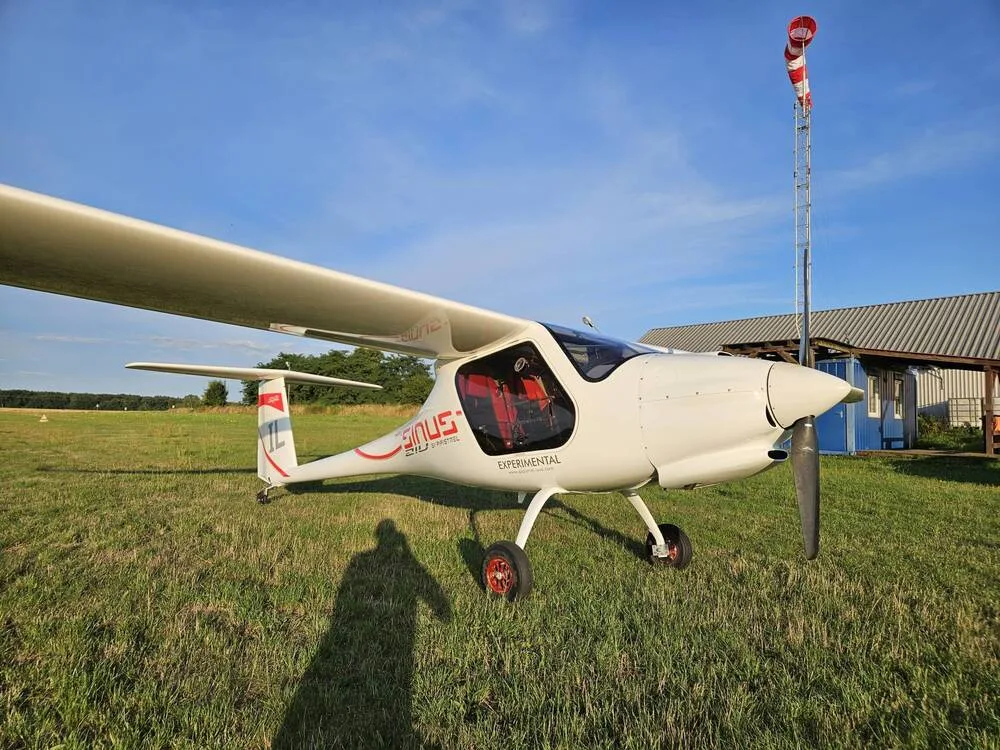 A white Pipistrel Sinus aircraft with red accents and "EXPERIMENTAL" text is parked on a grassy airfield with a windsock.