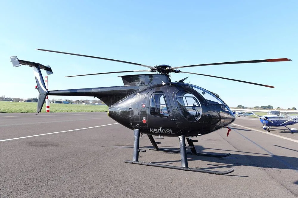 A black MD 500 helicopter (N500SL) is parked on an airfield tarmac, with other small aircraft in the distance.