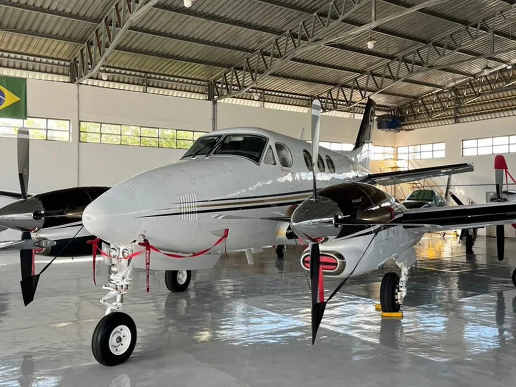 Front view of a white and black Beechcraft King Air 350 with gold stripes, parked inside a hangar with a Brazilian flag visible.