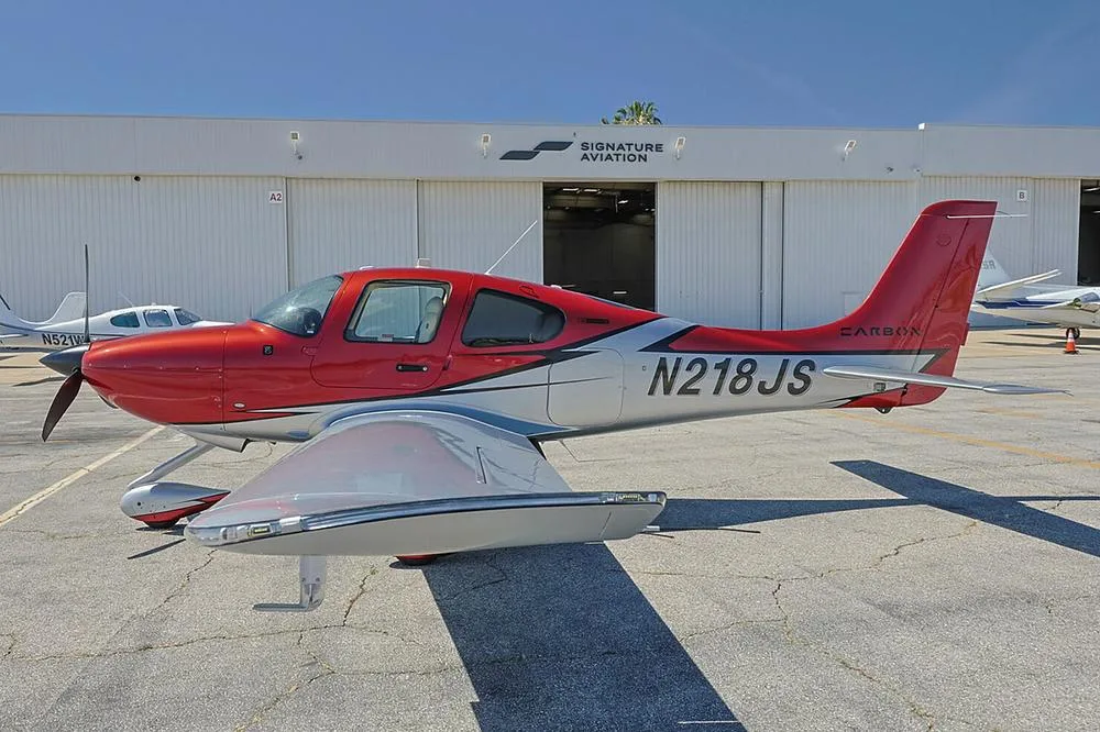 A red and silver Cirrus SR22 aircraft with registration N218JS parked on a tarmac in front of a Signature Aviation hangar.