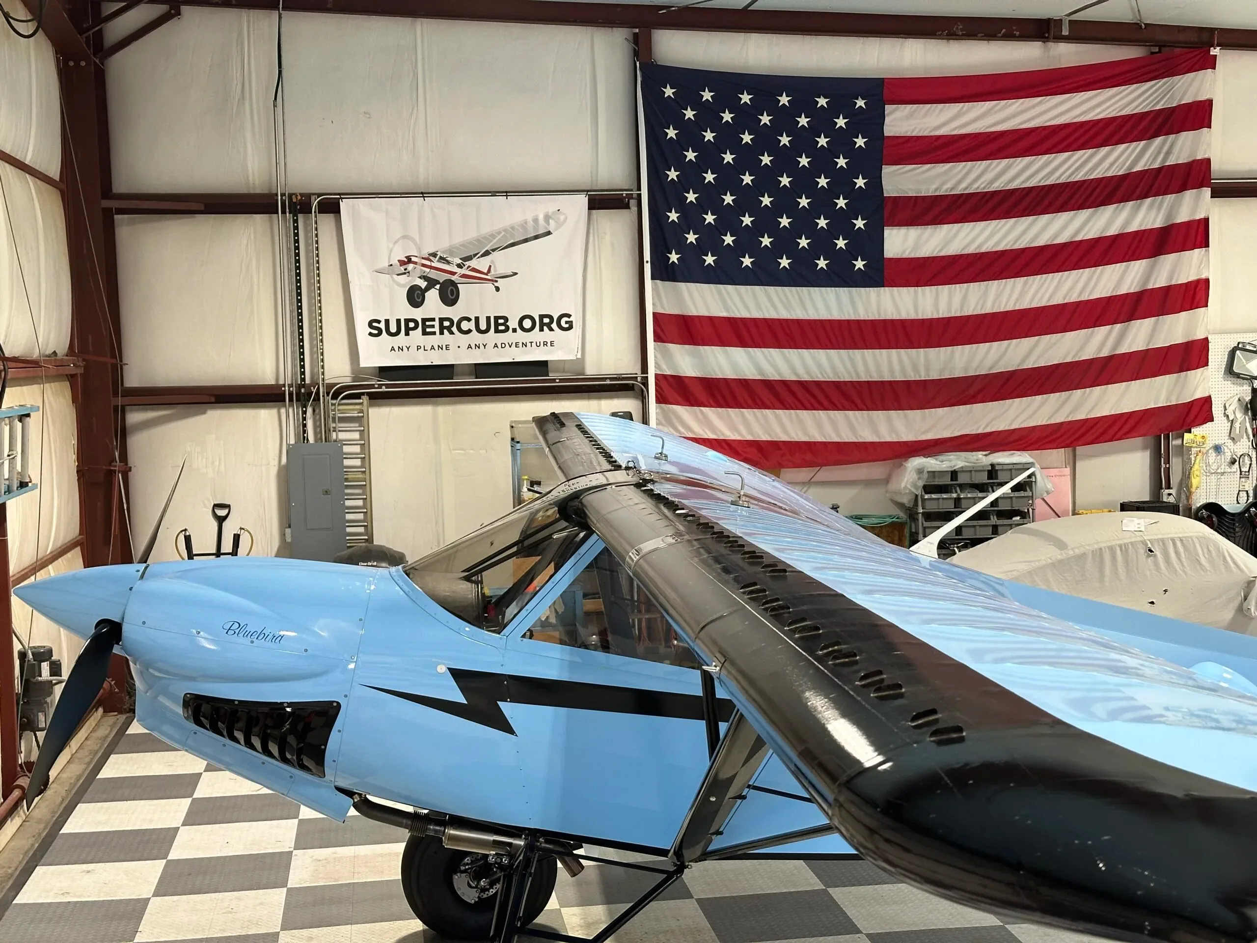 A light blue "Bluebird" aircraft with black wing struts and large tires, parked in a hangar with an American flag and a "SUPERCUB.ORG" banner.