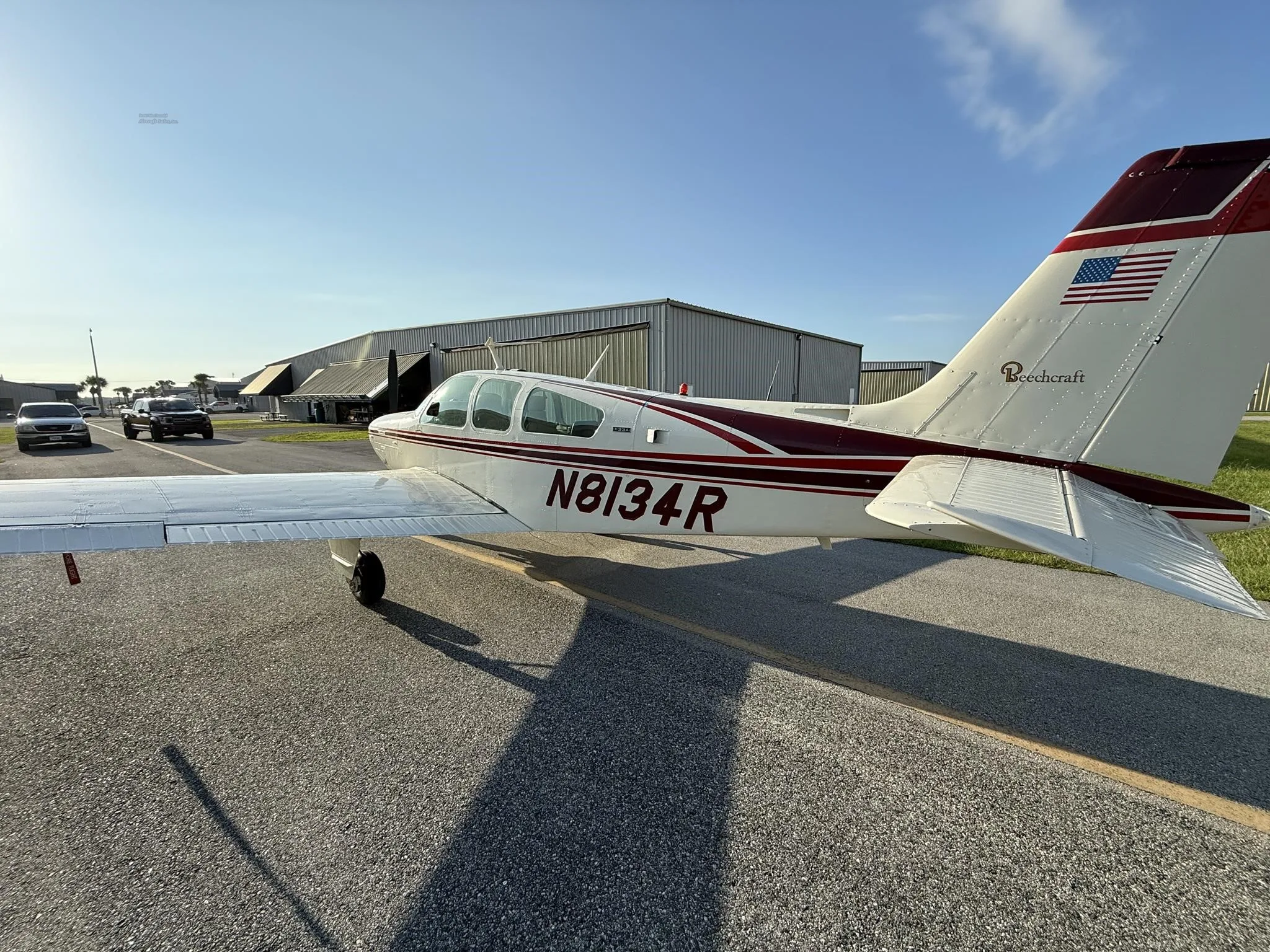 A white and maroon Beechcraft Bonanza N8134R is parked on an airfield with hangars in the background under a sunny sky.