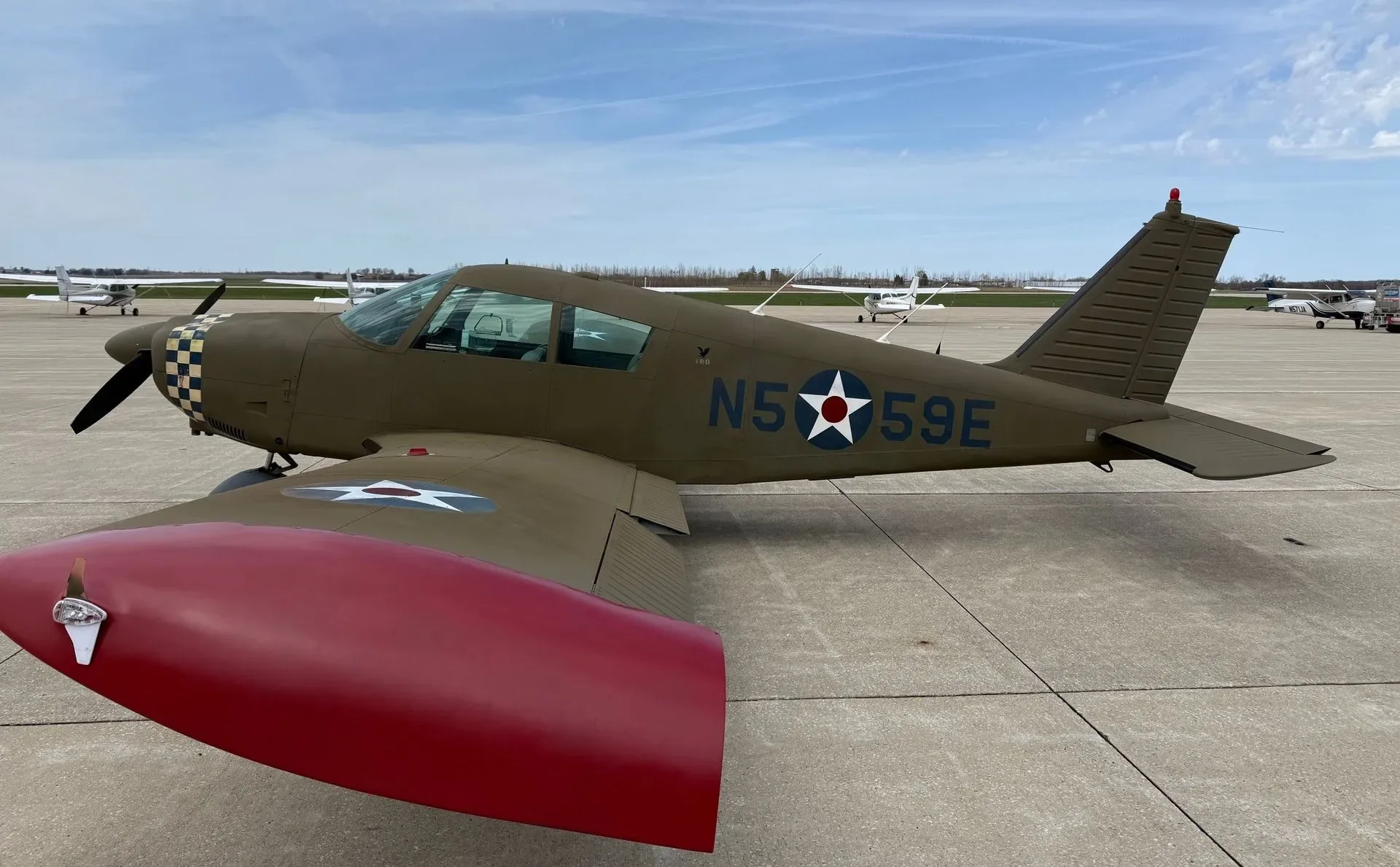 A single-engine aircraft with olive drab paint, checkered nose, red wingtips, and US military star insignia parked on an airfield.