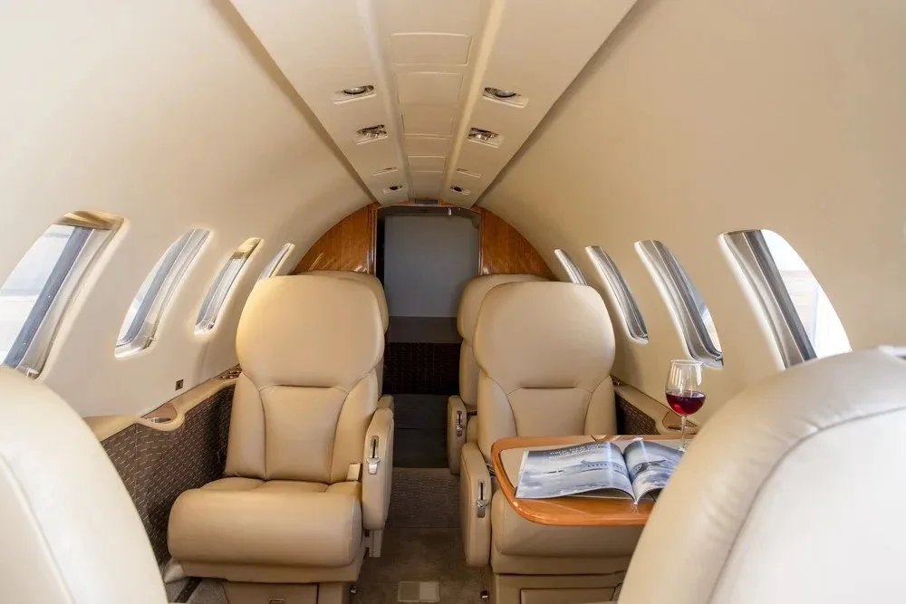 The interior cabin of a private jet with light tan leather seats, a small table with a magazine and a glass of red wine, viewed from the rear.
